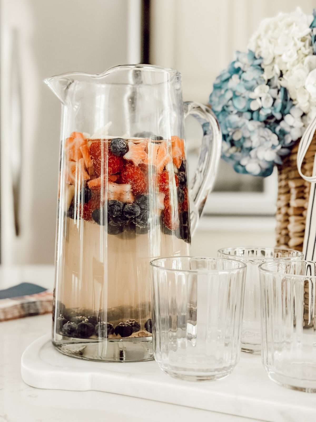 Glass pitcher of white wine sangria with vodka on kitchen counter with glass cups and a basket of flowers.