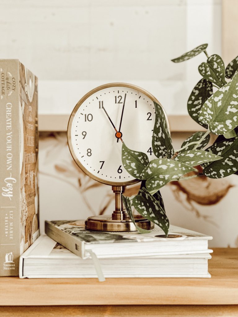 Vintage style pedestal clock on an old desk with books and a faux plant. 
