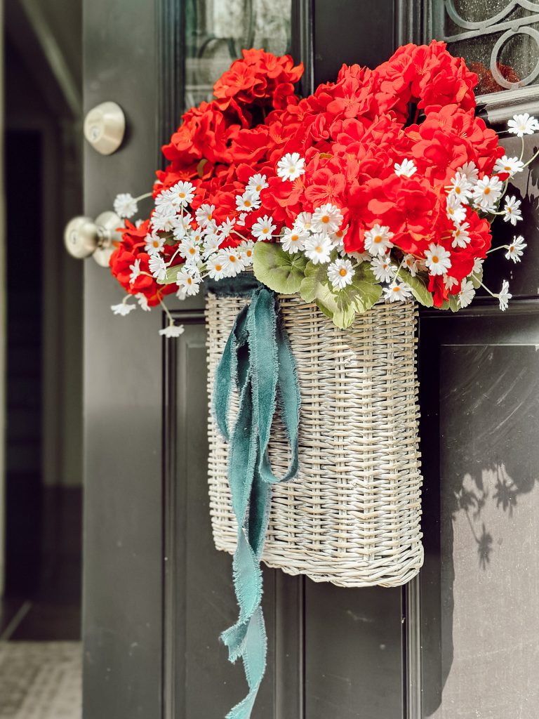 Front door basket with red flowers, white daisies, and a blue ribbon.