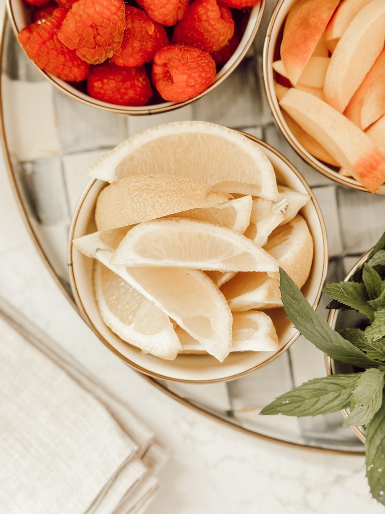 Small pinch bowls of sliced lemons, fresh raspberries, and sliced peaches for garnishes in lemonade.