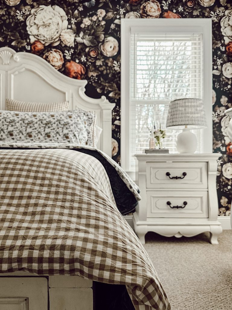 Gingham bedding on a king size bed with a large floral wallpaper accent wall behind it, and a nightstand with a lamp and fresh flowers.