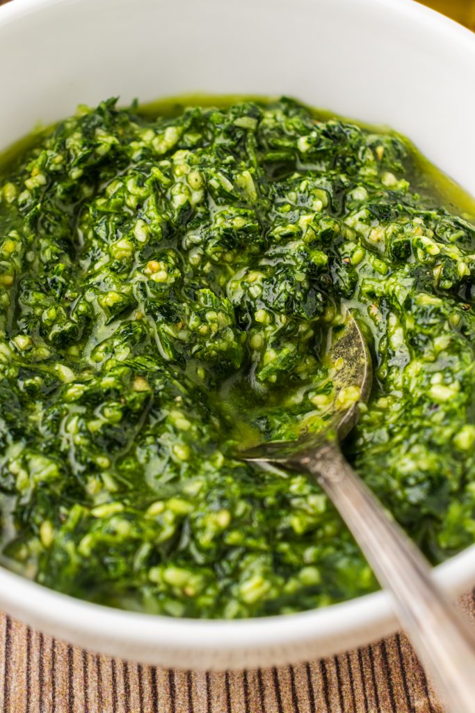 White bowl of fresh parsley pesto with a silver spoon in it for mixing into pasta dishes.