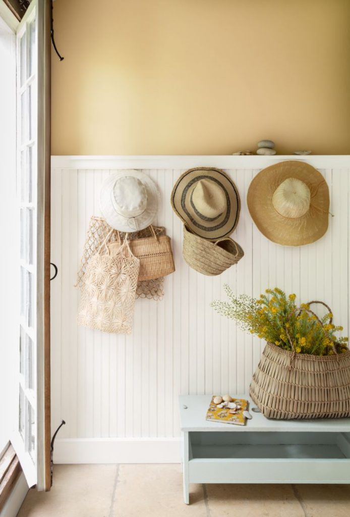 Country entryway with white beadboard and yellow walls. Straw hats hanging on pegs and a bench with a basket of flowers.