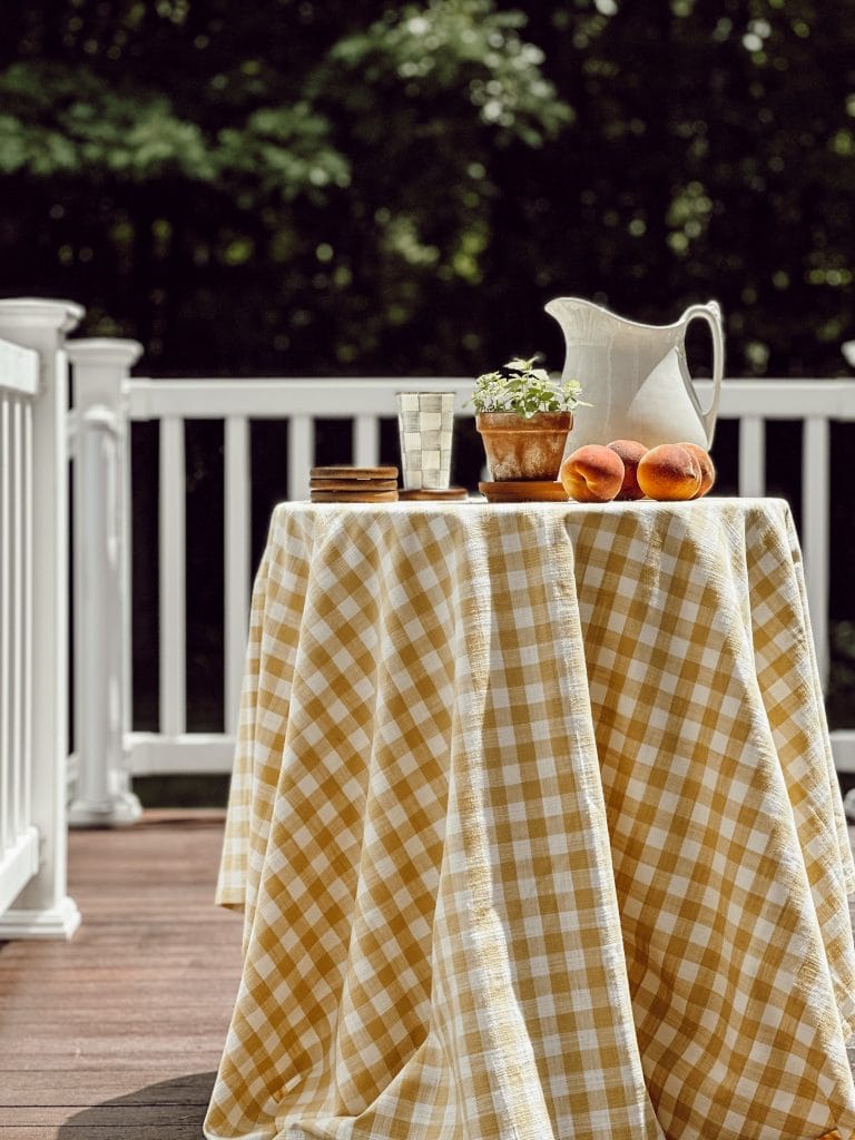 Decorating with butter yellow ideas includes this gingham tablecloth on a table with a white pitcher, peaches, and potted mint.