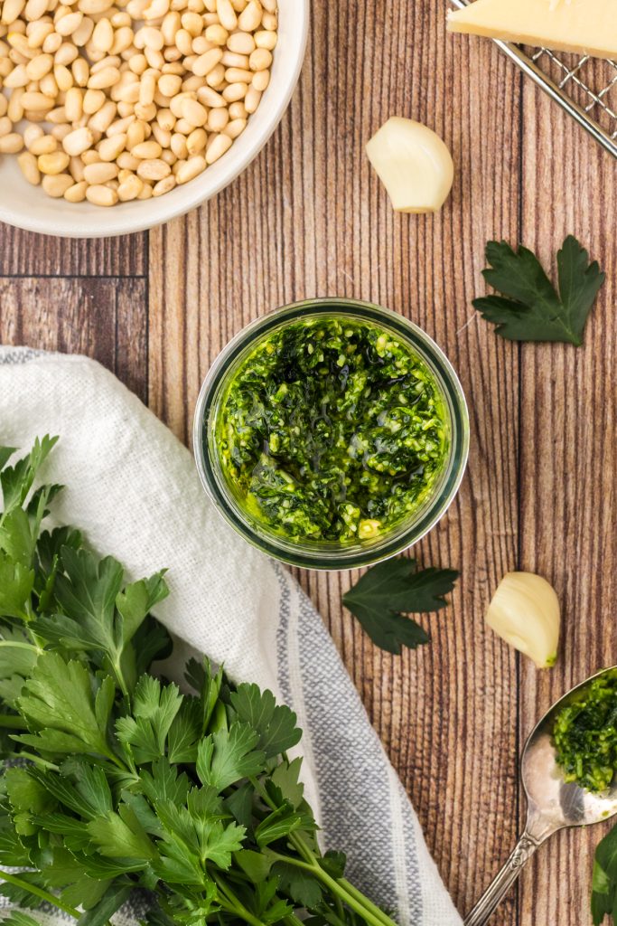Mason jar with parsley pesto on a wooden table with garlic cloves, pine nuts, and bunches of parsley around it.