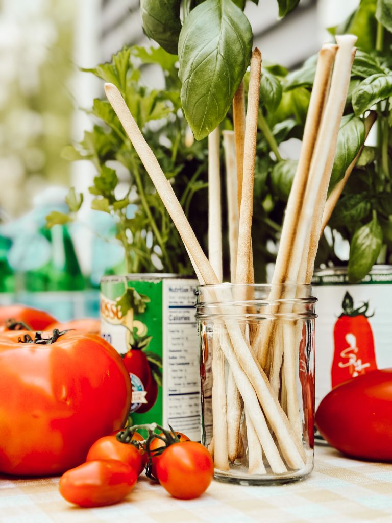 Outdoor table with gingham tablecloth, mason jar with breadsticks, fresh tomatoes, and basil for al fresco dining.
