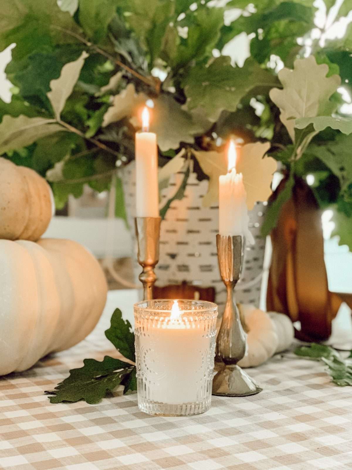 Fall vignette with candles, pumpkins, and oak branches in an olive bucket on a gingham tablecloth.