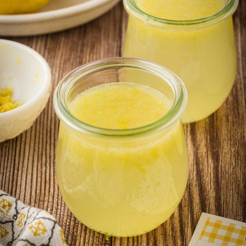 Glass jars of lemonade concentrate with a bowl full of lemons, kitchen towel, and gift tags, on a wooden surface.