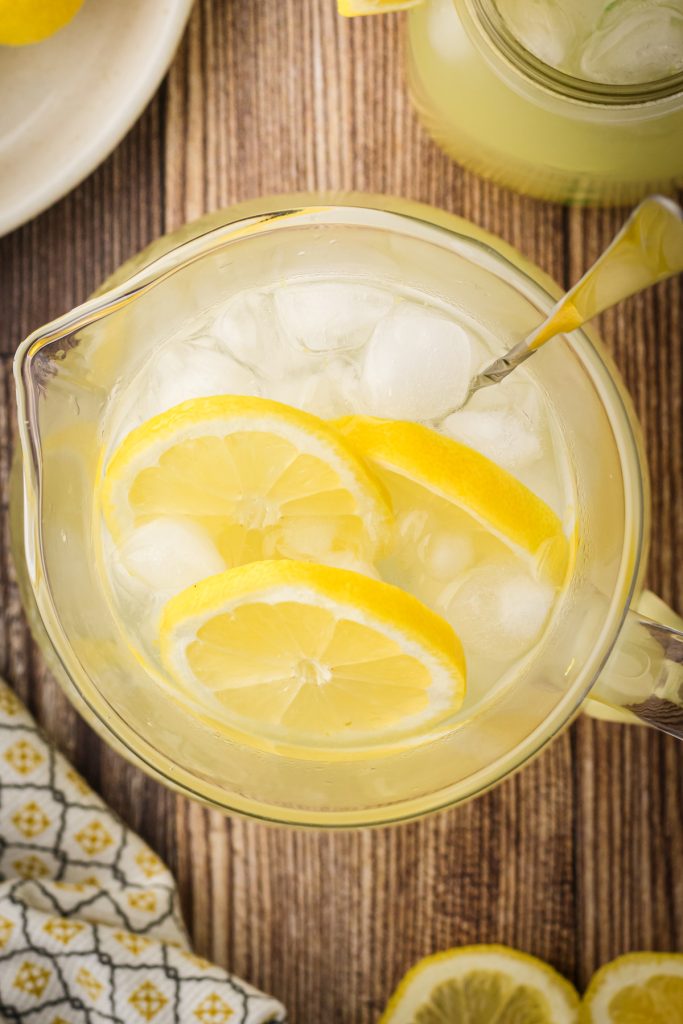 Glass pitcher of homemade lemonade on a wooden table with fresh lemon slices and ice cubes.