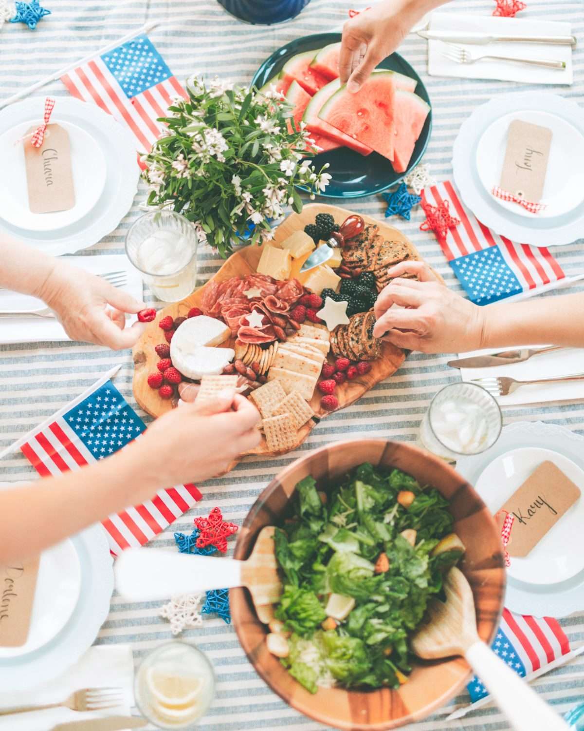 4th of July foods for a summer cookout include a charcuterie board, salad, sliced watermelon, and small American flags scattered on a table.