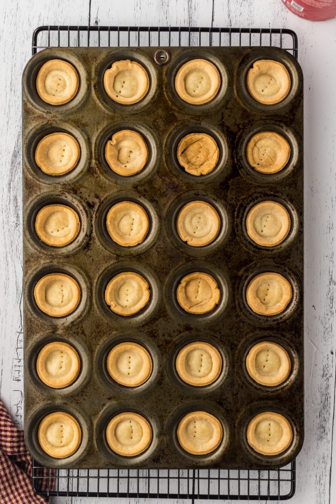 Baked pie crusts in mini muffin pan on a cooling rack on a wooden surface. 