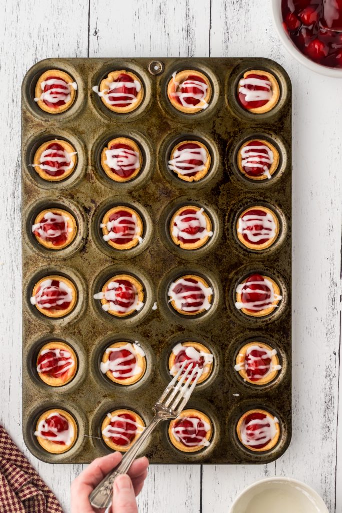 Drizzling the sugar glaze over the top of the mini cherry pies with a fork.