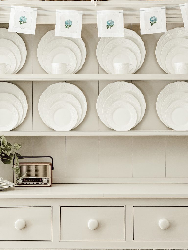 Kitchen hutch filled with white dishes with a hydrangea coaster garland hanging on the top.