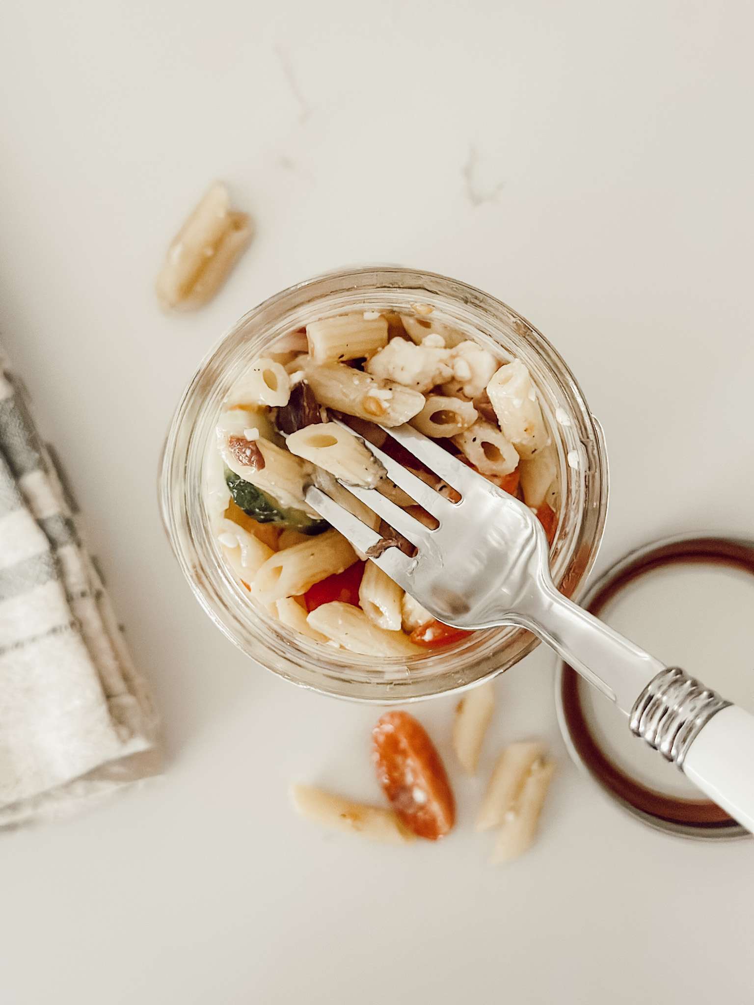 Mediterranean penne pasta salad in a mason jar with a fork for easy eating a summer barbecue.