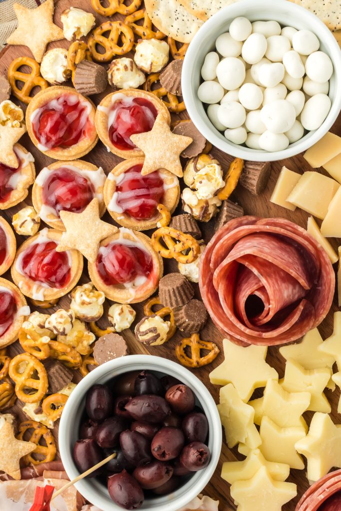 Sweet and savory ingredients on a wooden board include olives, yogurt covered raisins, salami, cheese, and mini cherry pies.