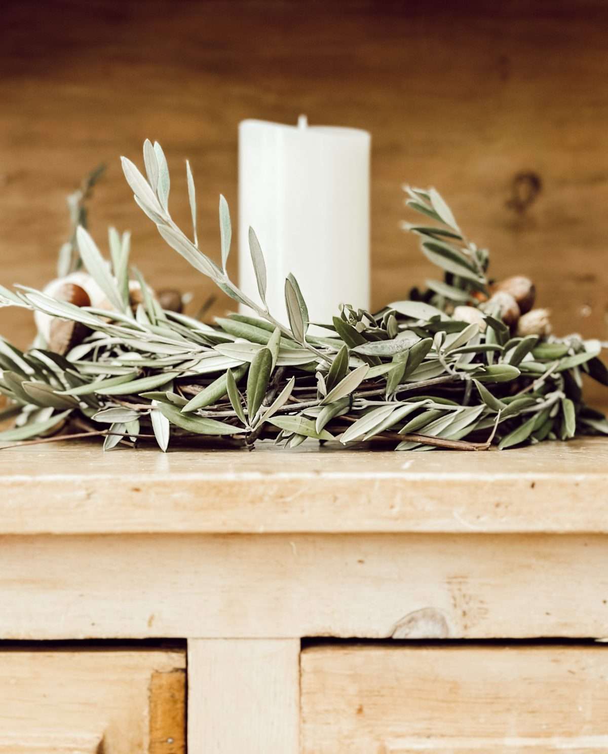 A DIY fall candle wreath made with olive branches, nuts, a white pumpkin, and a pillar candle in the center.