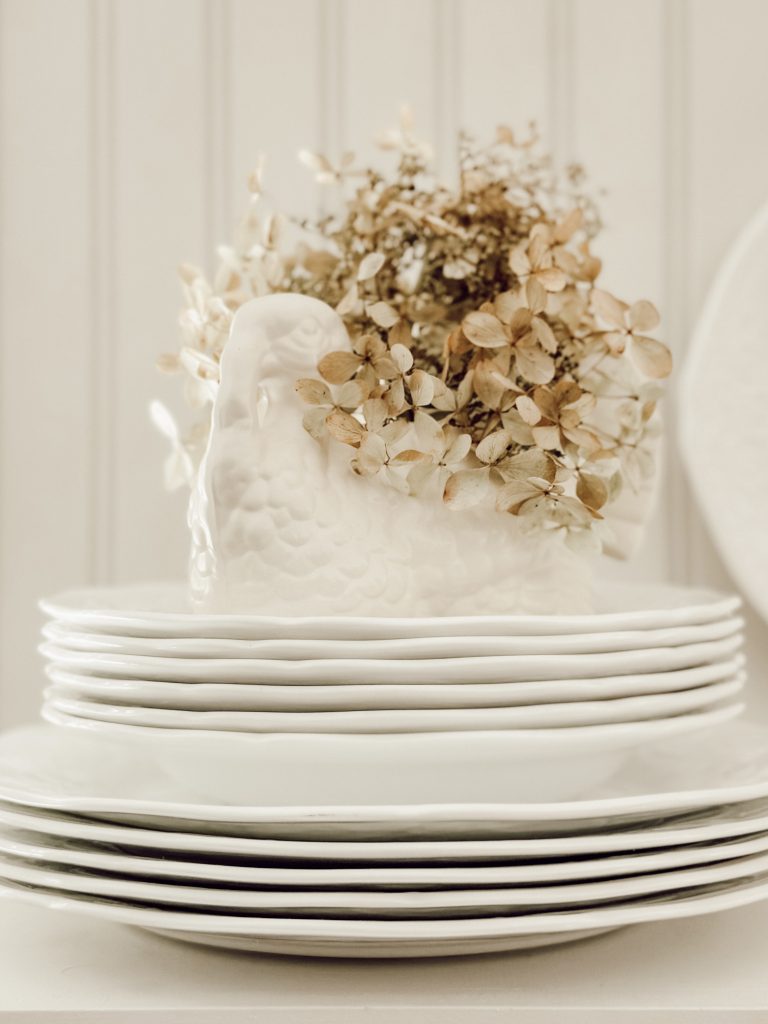 Dried hydrangea blooms in a turkey vase on top of stacked white plates for cozy fall decorations.