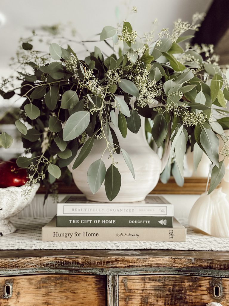 A vase full of seeded eucalyptus on a stack of books with a bowl of apples and candle for cozy home decor.