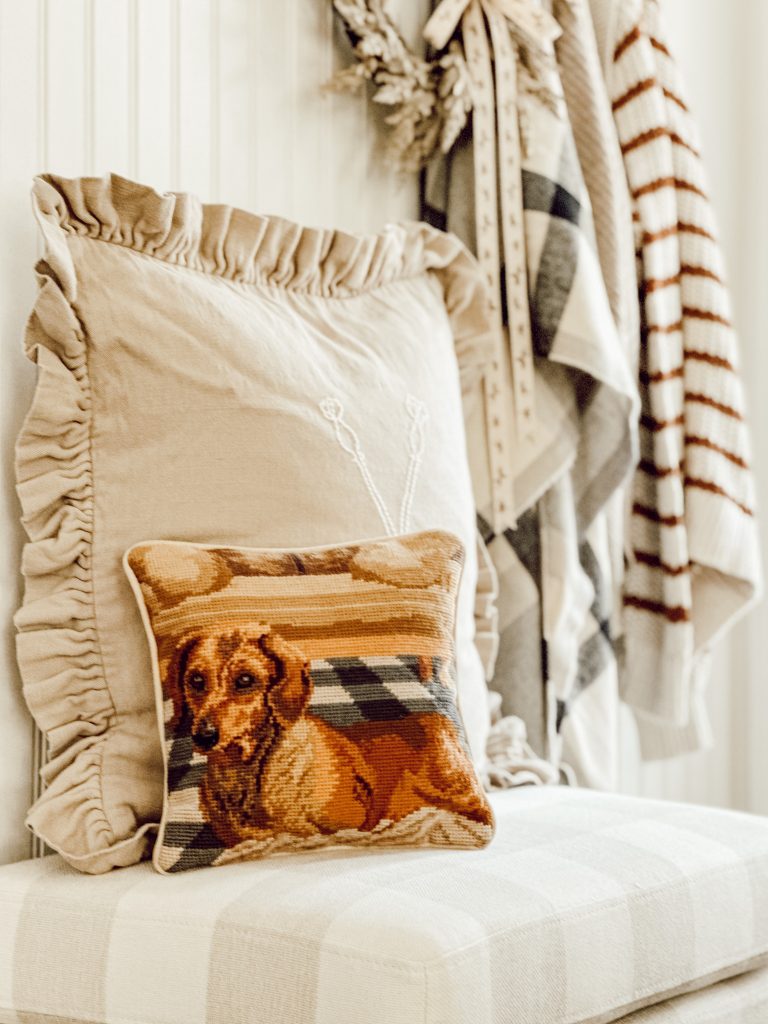 Neutral fall decorations in entryway for the fall season with a buffalo check ottoman, throw pillows, wreath, and sweaters.
