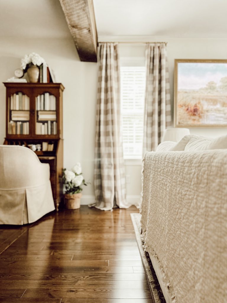 Neutral living room decorated for fall with a quilt draped on the back of the couch and buffalo check curtains hanging on the windows.