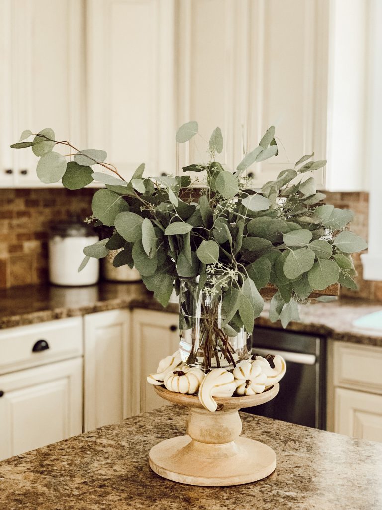 Vase of silver dollar eucalyptus with white mini pumpkins on kitchen counter.