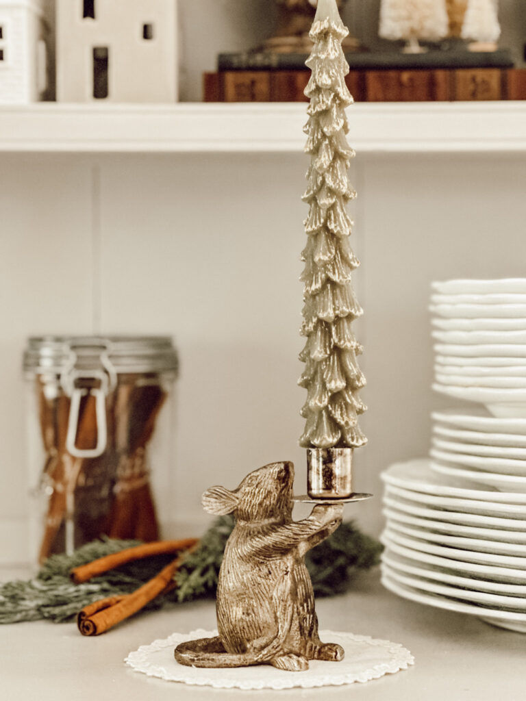 Christmas vignette on kitchen hutch with a brass candleholder, tree candle, glass jar of cinnamon sticks, and a stack of plates.