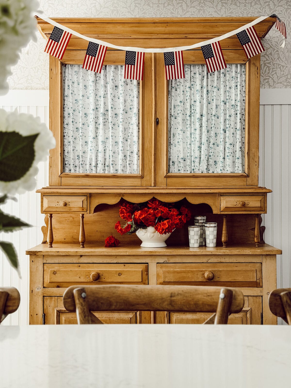 DIY American flag garland hanging on a wooden hutch with red geraniums and checkered cups.