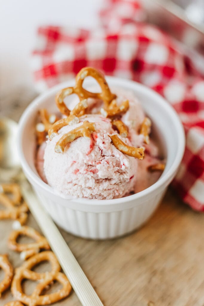 Small white bowl of ice cream topped with chopped pretzels next to gingham towel.