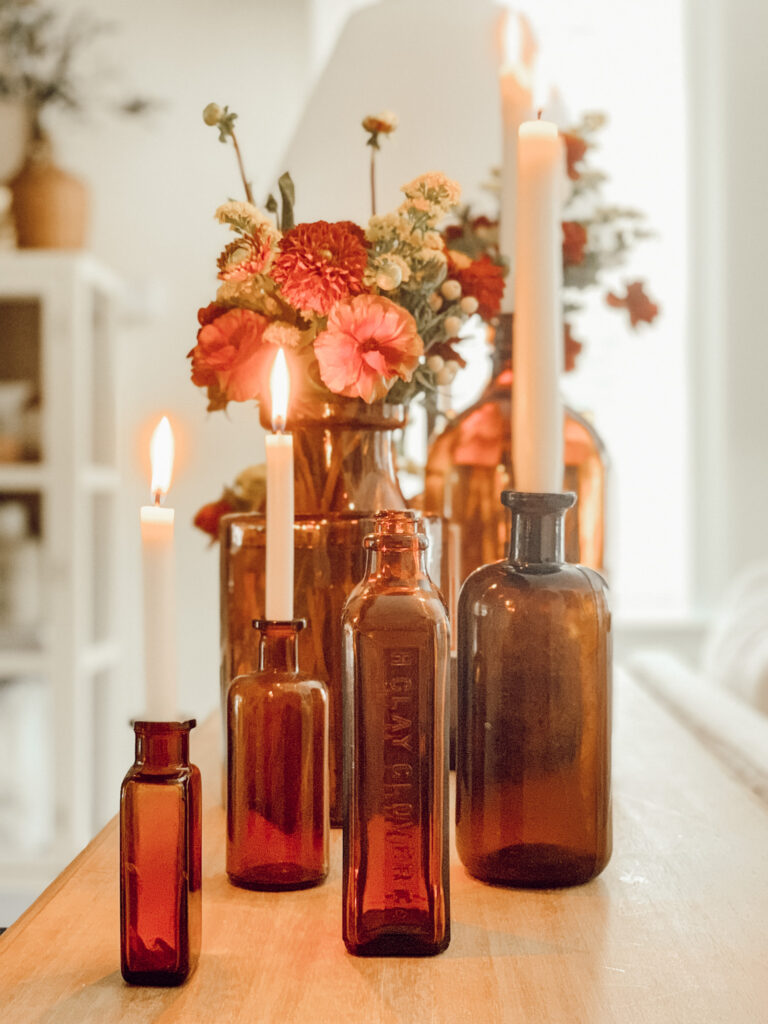 Amber glass bottles filled with fall flowers and candles on a wood entryway table.