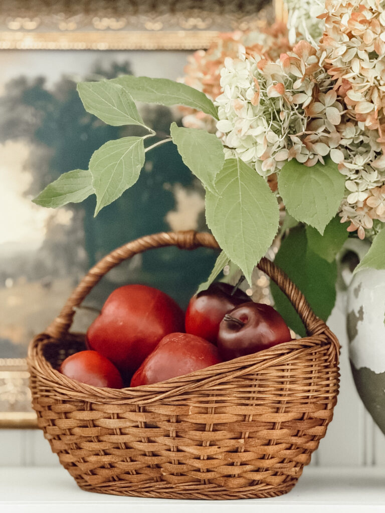 Hydrangea stems in a vase with a basket of red apples for cozy fall decorating.