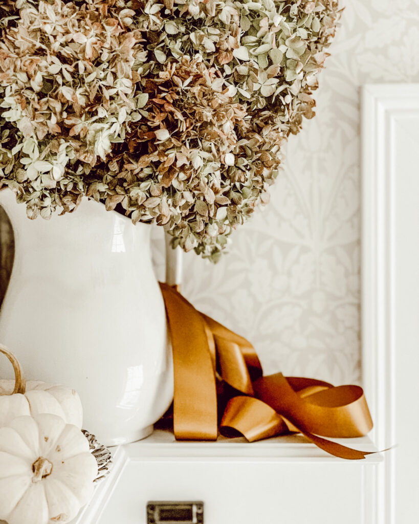 Ironstone pitcher filled with hydrangea flowers and tied with a gold satin bow and white pumpkins next to it.